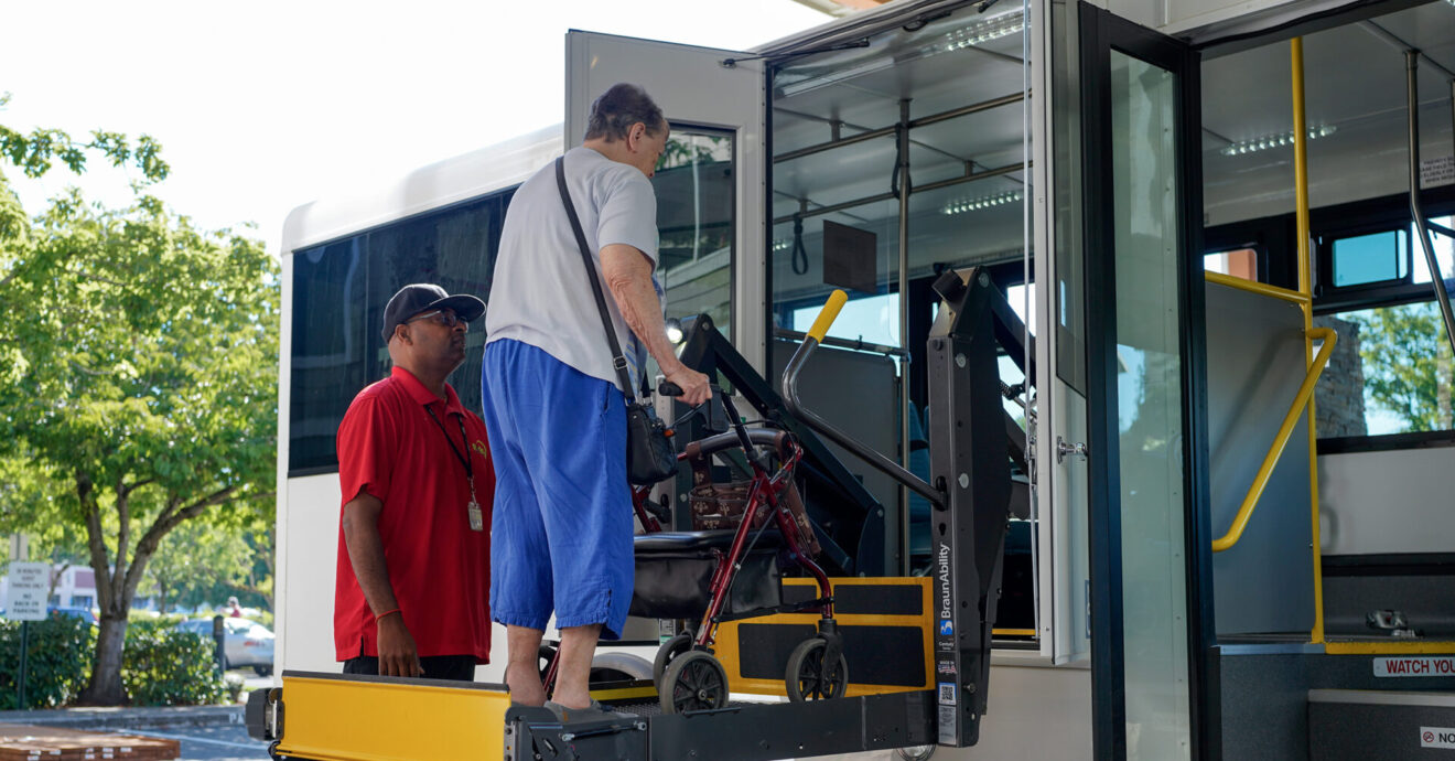 A black male driver is assisting a woman with a mobility device off of a bus