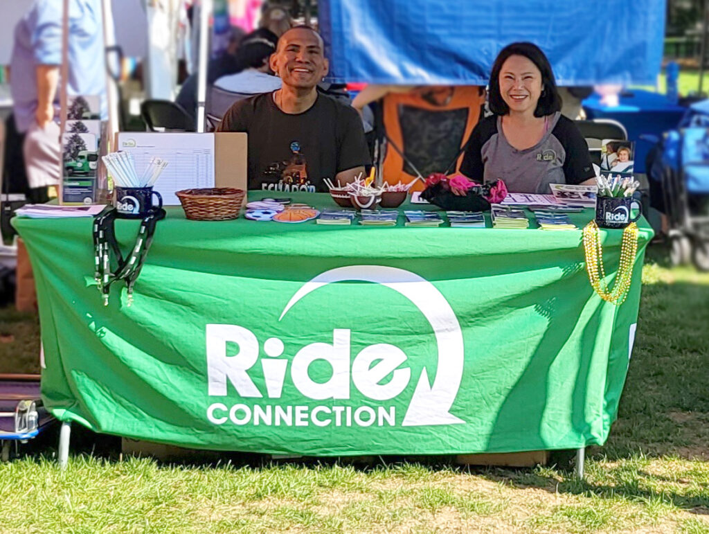 Two Ride Connection employees, a Hispanic man and an Asian woman, at a tabling event outdoors