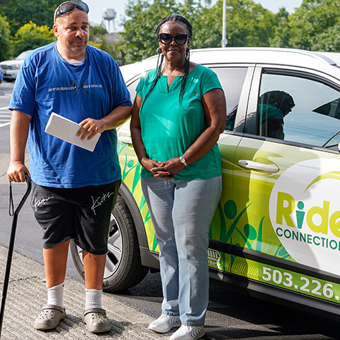 Ride Connection driver, a black woman and a male customer using a walking cane pose in front of an Electric Vehicle