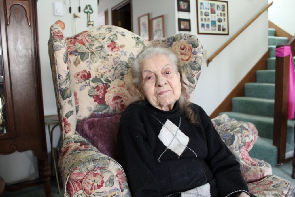 An older Lebanese woman, Claire Dallal sits on a floral couch in her home.