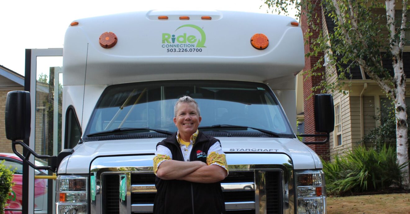 A male driver stands with his arms crossed over his chest. He is standing in front of a Ride Connection shuttle.
