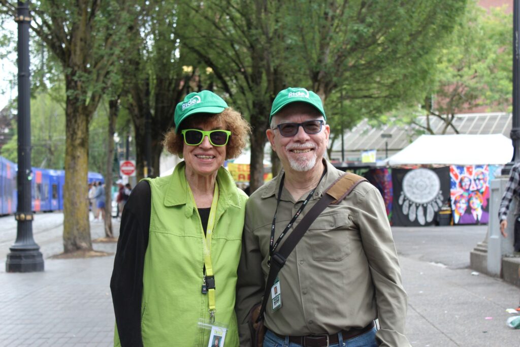 Two volunteers, a man and a woman, stand together wearing Ride Connection hats.